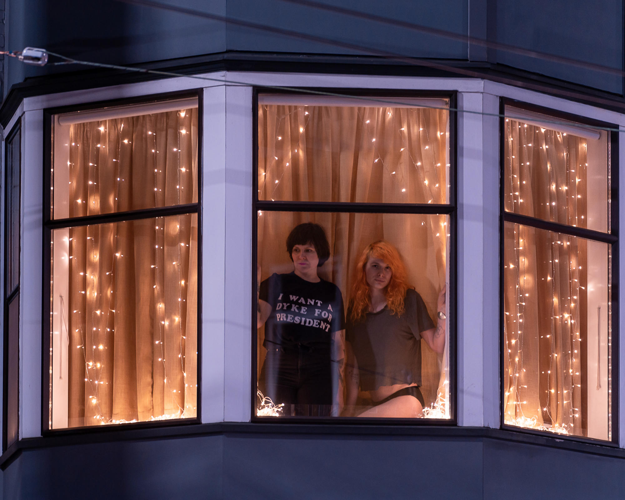 Two people in their window, one wearing a T-shirt that says "I want a dyke for president."