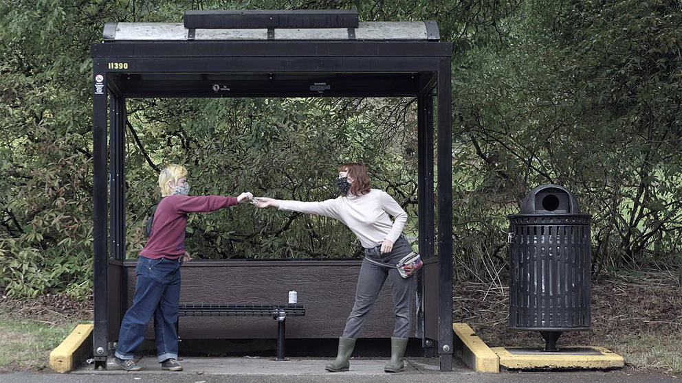Two people in front of a bus stop, handing over hand sanitizer