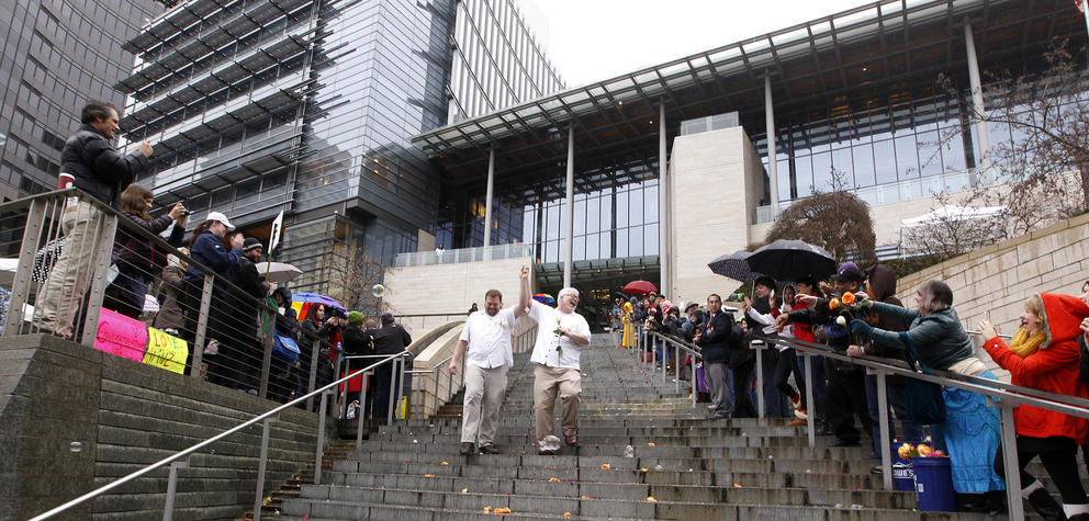Two men wearing white raise their hands together as they walk down the steps of Seattle City hall. Onlookers flank each side. 