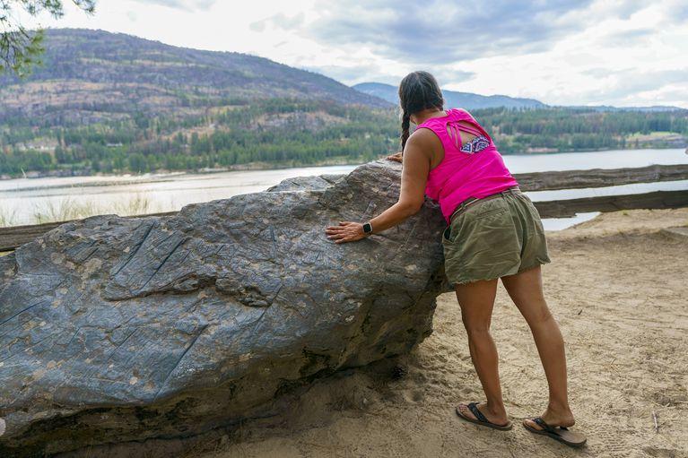 A woman stands next to a large stone overlooking a river