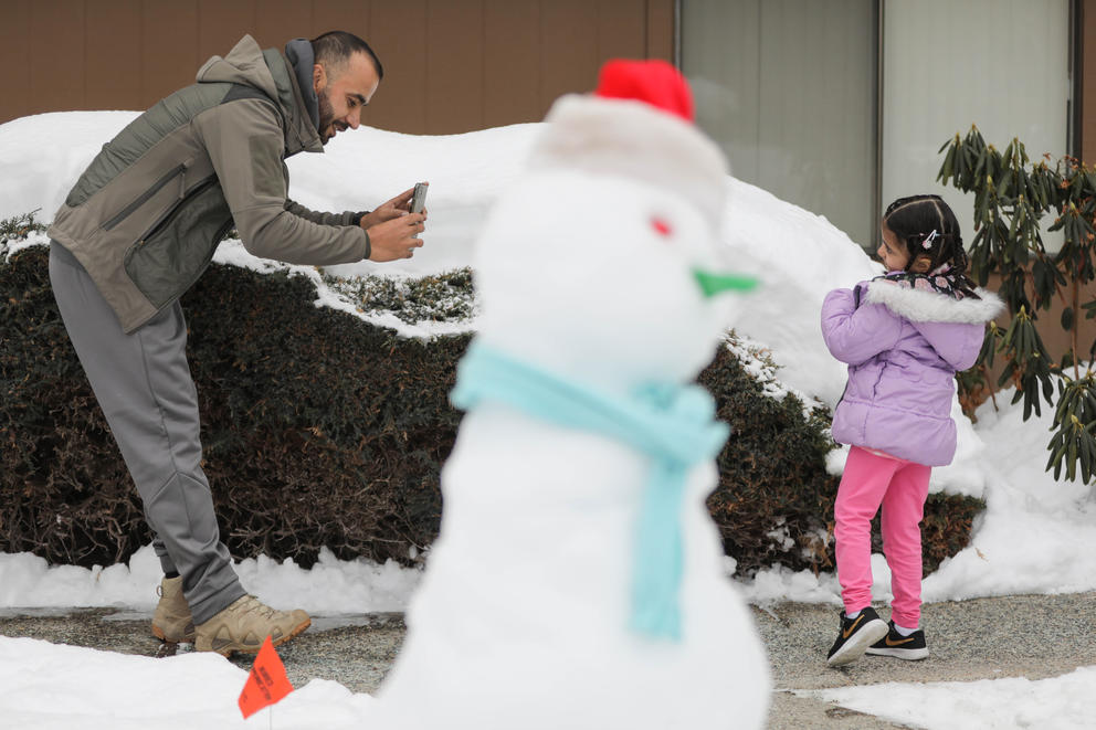 Reshad Al Noori takes a photo of his daughter Maryan, 4, outside their apartment. A snowman wearing a blue scarf and red hat is seen in the foreground.