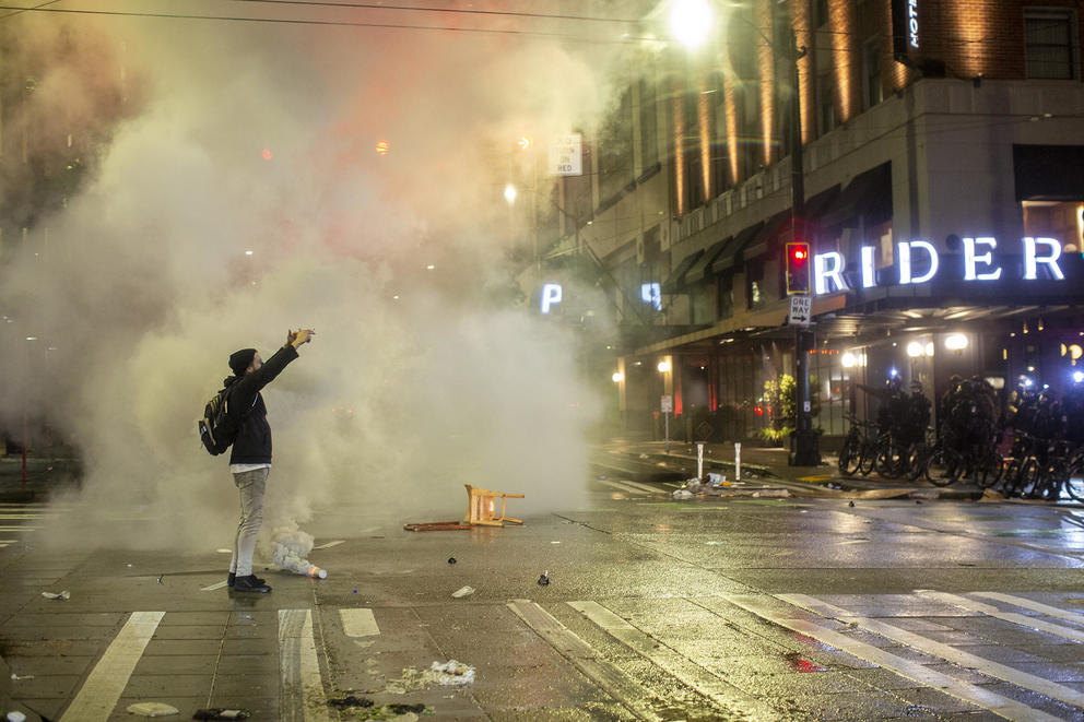 A man in a jacket and backpack stands in the middle of an intersection with a smoke canister at his feet. He has his arms raised above his head, pointing at police officer on bicycles during a Seattle anti-racism protest.