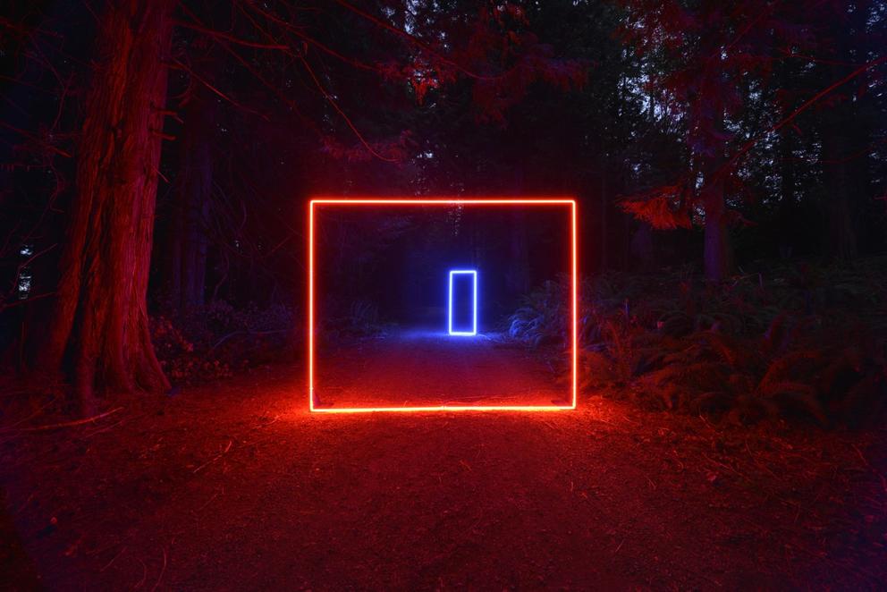 A quare of red neon lights up a wooded backroad. In the distance, you can see another neon sculpture, a blue rectangle standing up.