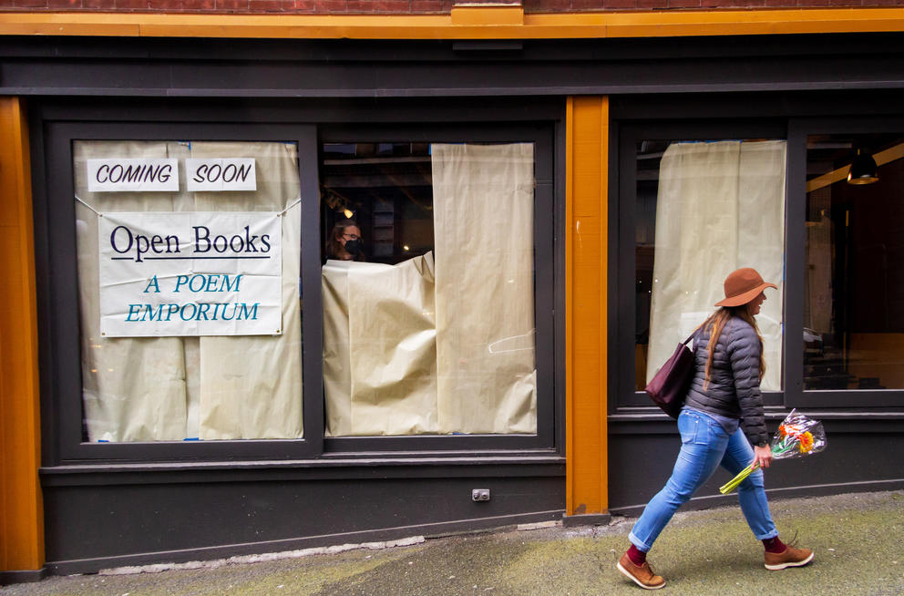 Person in orange hat and flowers in hand walks by window that is still covered in paper. Behind the window, a person is taking the paper off. To her left, a sign reads: Open Books: A Poem Emporium