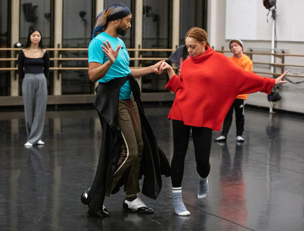 Person in blue t-shirt and black pants holds the hands of a person in black pants and red sweater as they are dancing and practicing steps in a rehearsal dance room