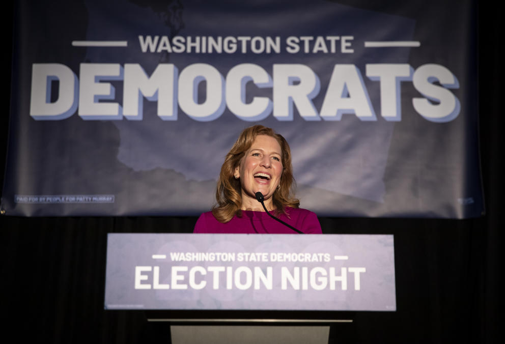 A woman stands behind a podium with a sign behind her that reads Washington State Democrats