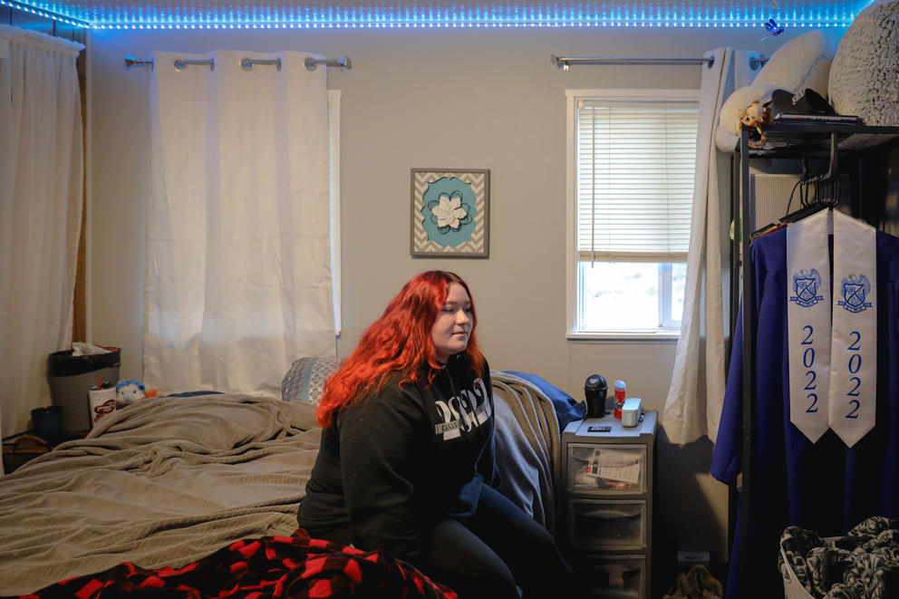 A young woman with red hair sits on her bed in her bedroom, a graduation gown with 2022 on it hangs on a rack at the right