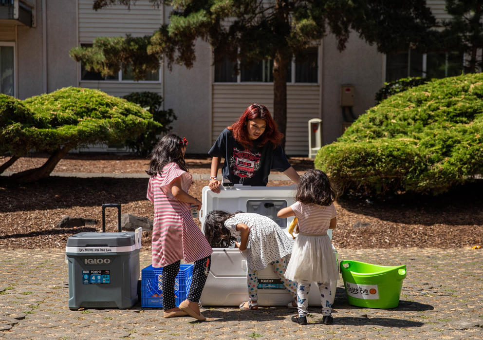 Three children in dresses reach into a cooler at an apartment building as a food bank employee assists.