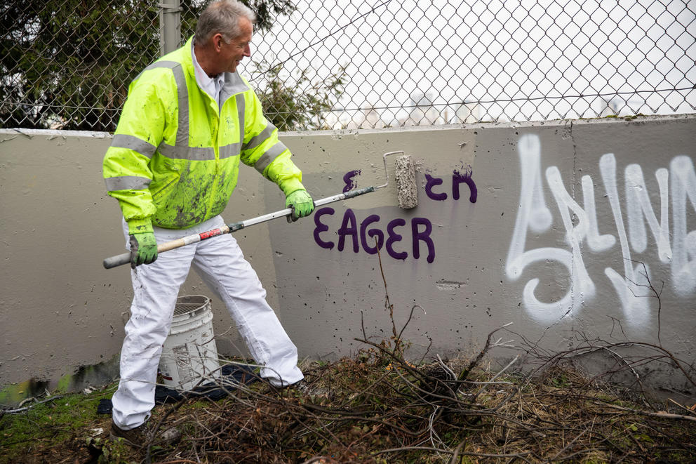 A man in white pants and a hi-vis vest paints over graffiti reading "eager"