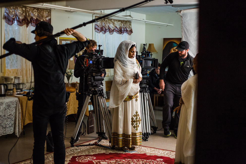 Person in white clothing in a room surrounded by a film crew holding up a mic