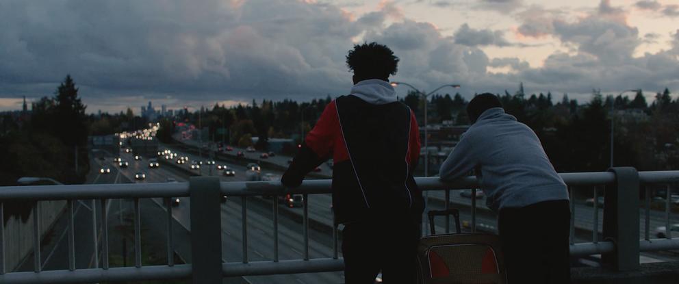 two people with their back to the camera, facing a highway, below it a highway and above it a darkening sky full of thick clouds