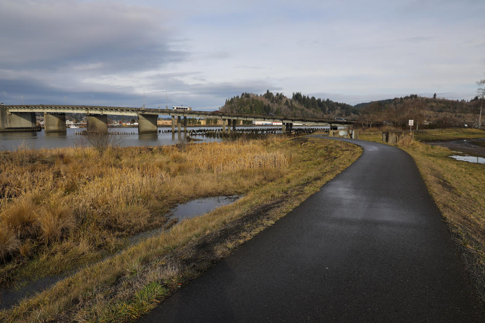 An asphalt walk way runs along a raised earthen berm, a river and bridge are seen in the background