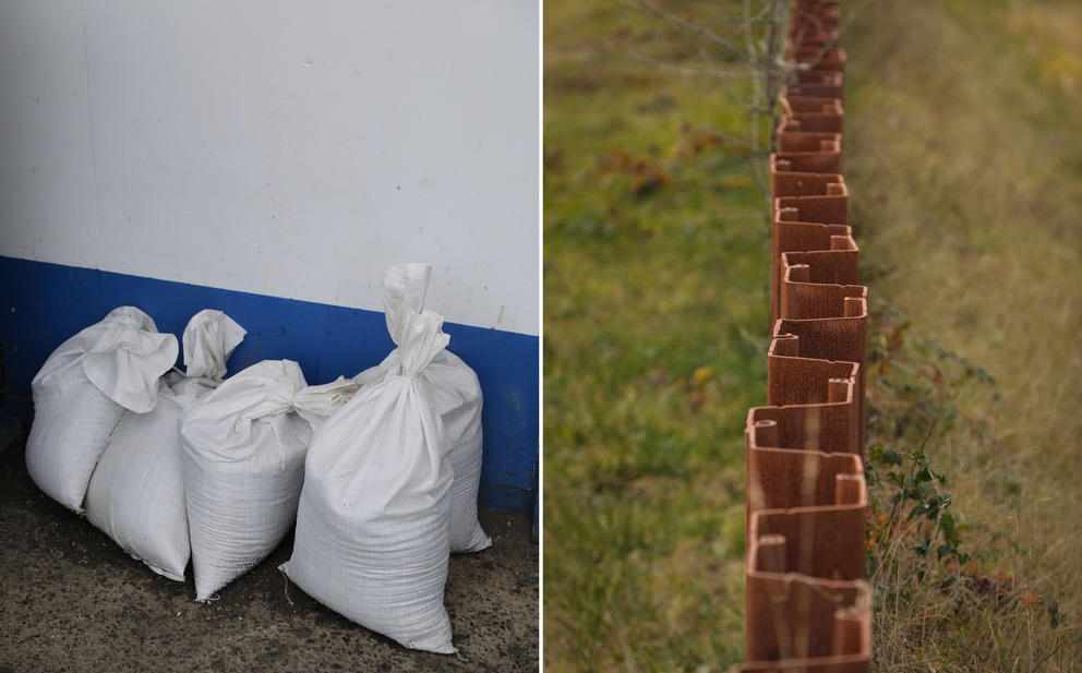 On the left, white bags filled with sand sit against a wall. On the right, a sheet metal levee wall splits the frame with grass on either side
