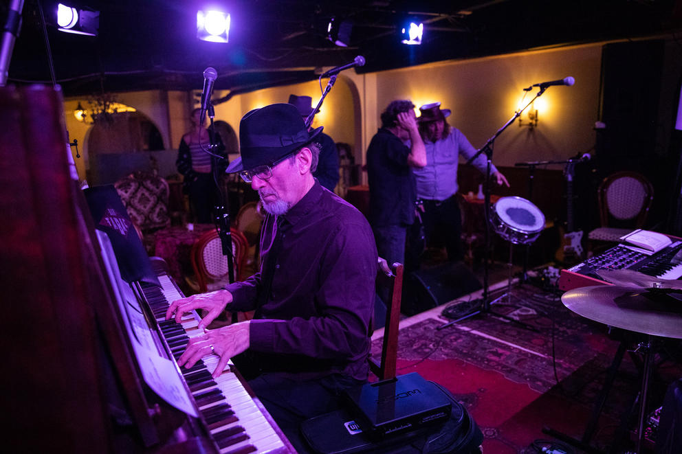 Man with a dark hat sits at a piano in what looks like an underground venue