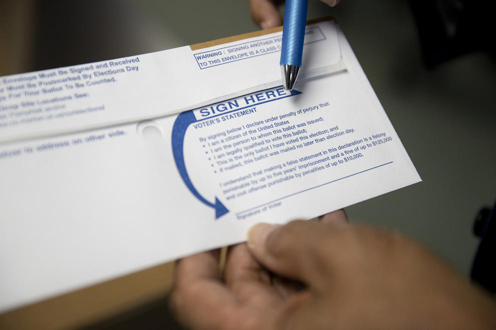 The tip of a pen points to a signature line on the back of a mail-in ballot
