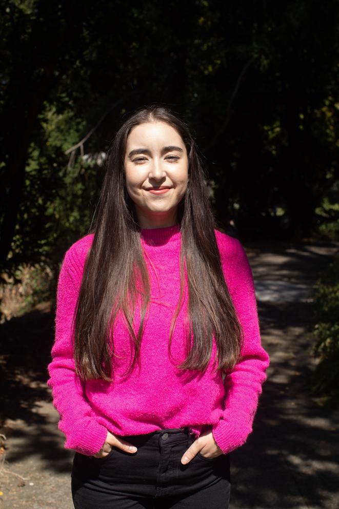 Margot Spindola looks straight at the camera, hands in her pockets, wearing a pink top