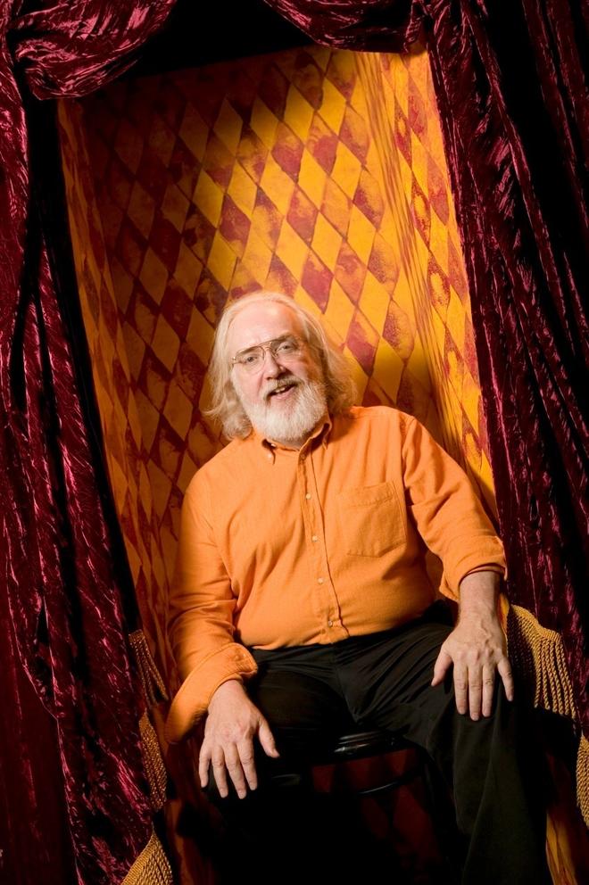 Man in orange shirt with grey beard and grey hair but bald top of head, orange circus pattern displayed behind him