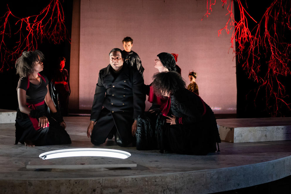 5 people sit in a semi-circle on a stage, they are cloaked in dark clothing and are looking at a hole in the ground with white light coming from below. The backdrop is washed red.