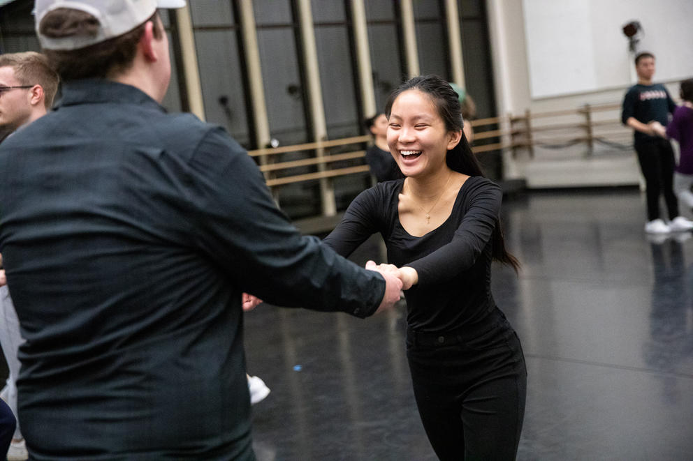Person holds the hands of another person, both dressed in black, as they are dancing together. The person on the left is smiling widely. They are in a rehearsal space.