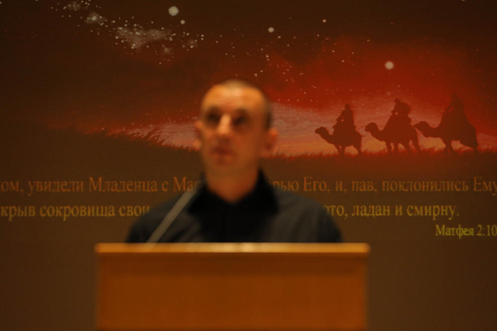 A out of focus man stands at a lectern, behind him a verse is writing in Cyrillic, above it a christmas scene with three wisemen on camels is projected
