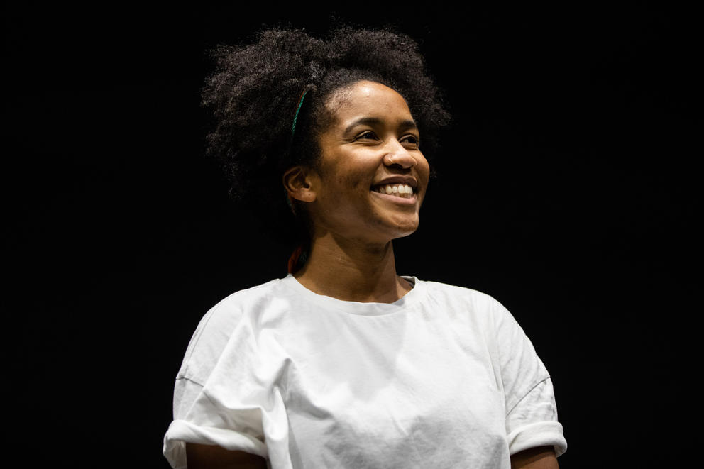 a woman in a white t-shirt smiles on a black-box theatre stage