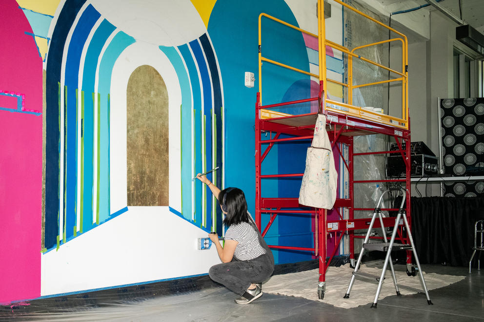 A woman paints a mural on a wall of arches of varying blues with a metallic gold center