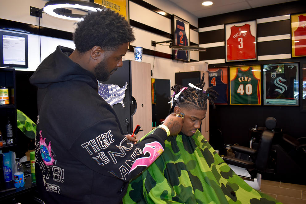A barber gives a seated client a haircut in a shop decorated with sports jerseys on the wall.