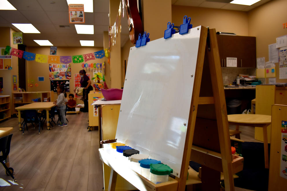 Children at a childcare/preschool play in the background watched by an adult, with an easel in the foreground