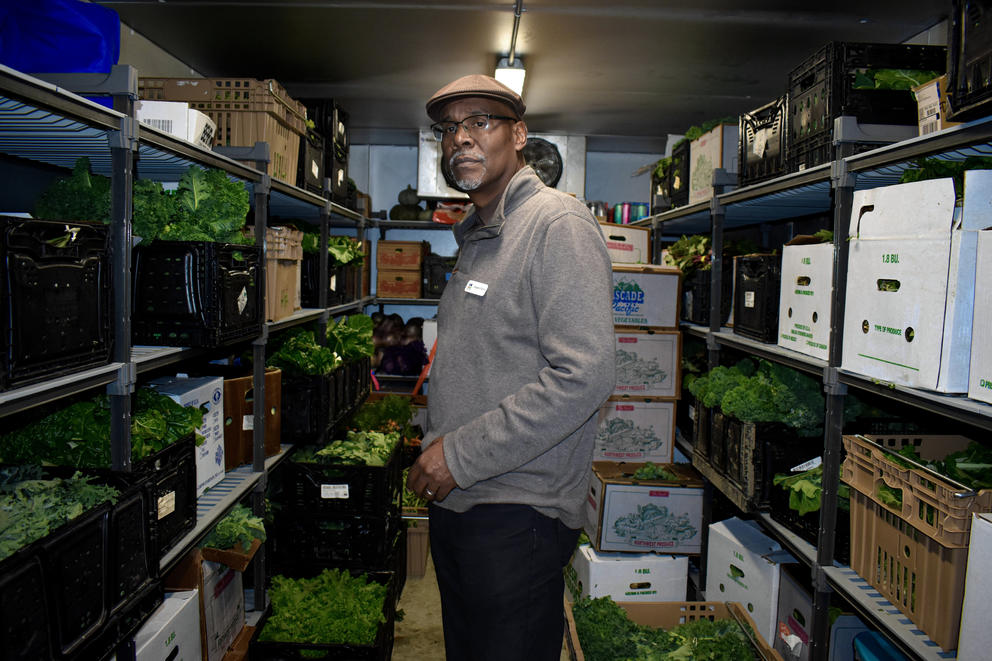 An adult in a gray sweater and a newspaperboy cap stands among produce in a walk-in fridge.