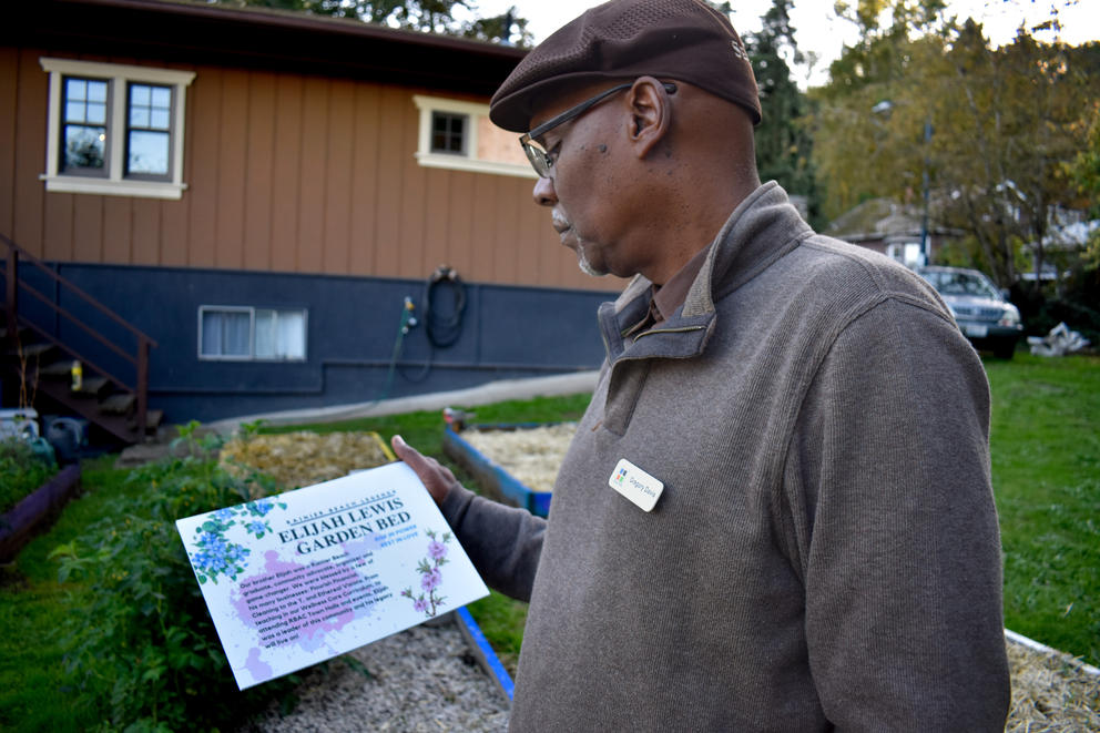 An adult wearing a sweater stands in a garden holding a garden sign that honors Elijah Lewis, a 23-year-old community organizer who was killed in 2023.