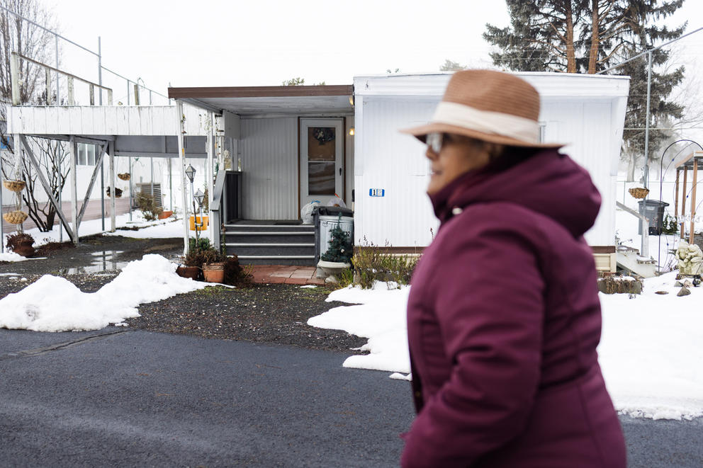 A woman walks through a Sun Tides mobile home park on a snowy day in January. She's wearing a big burgundy puffer jacket, paired with a straw hat on her head.