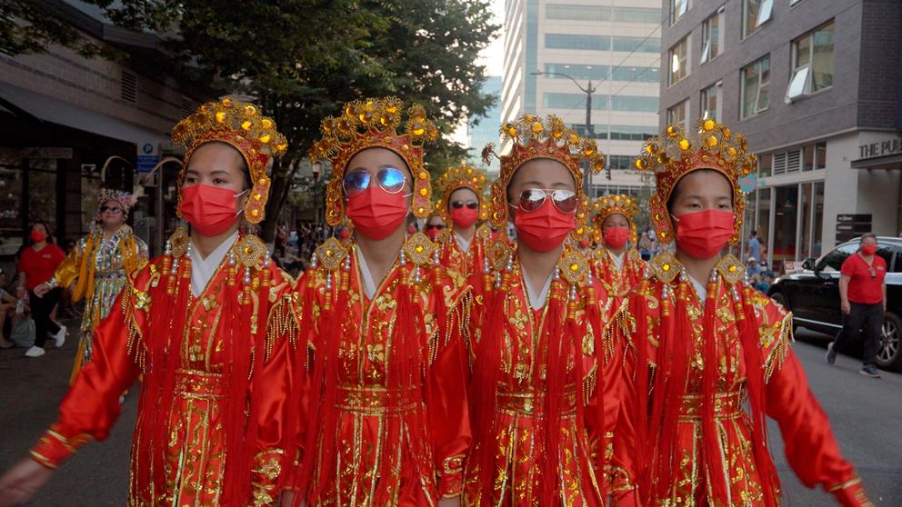 four young girls march in a parade wearing elaborate red and gold costumes, crowns and sunglasses