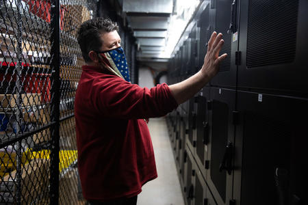 IT Construction Coordinator Michael Moore stands in a red shirt and mask, gesturing at a row of electronics at the Grays Harbor Public Utility District building in Aberdeen.