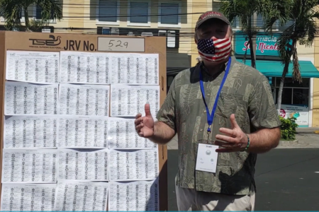 Doug Ericksen stands in front of a poster talking about the voting process in El Salvador, with palm trees and buildings behind him