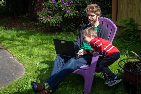Ryan Webber sits outside in a chair on the grass, working on a laptop as his son leans over his lap to look at the screen.