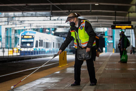 A wide shot of Lynn Chase, wearing a neon safety vest and black visor, following a tactile strip with cane at the Intl. District / Chinatown light rail station as a train arrives on the opposite platform.