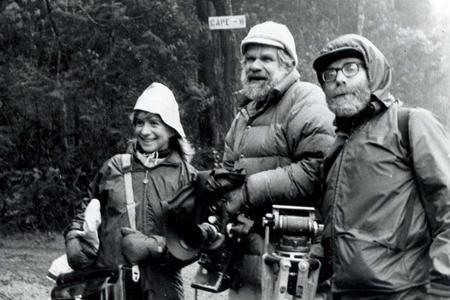 Black and white photo of three people in rain gear, a woman on the left, two men to the right of her