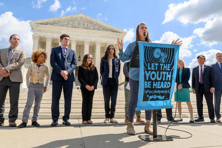 A group of youth plaintiffs stand in front of the Supreme Courthouse in suits, with one young woman giving a speech behind a teal poster.