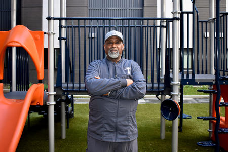 An adult in a grey tracksuit and a sports cap looks directly at the camera while standing in a play area.