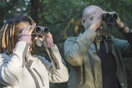 Black woman and Black man look through binoculars in wooded area