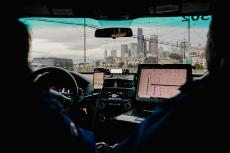 From the point of view in the back of a police car, a police officer sits in the driver's seat and looks at a laptop with the Seattle skyline in the windshield. 