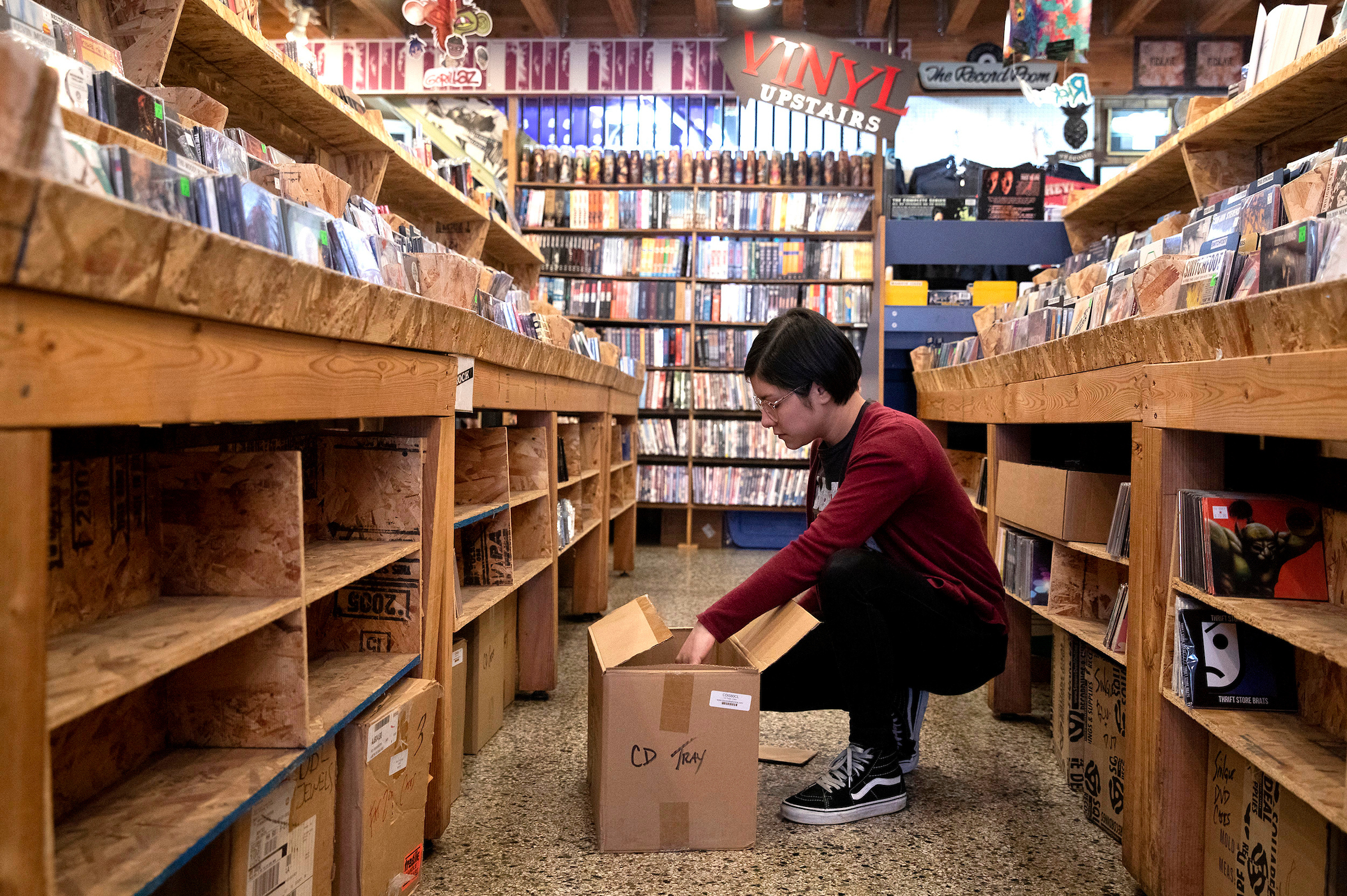 Person crouches with box of records in the middle of record store isle.