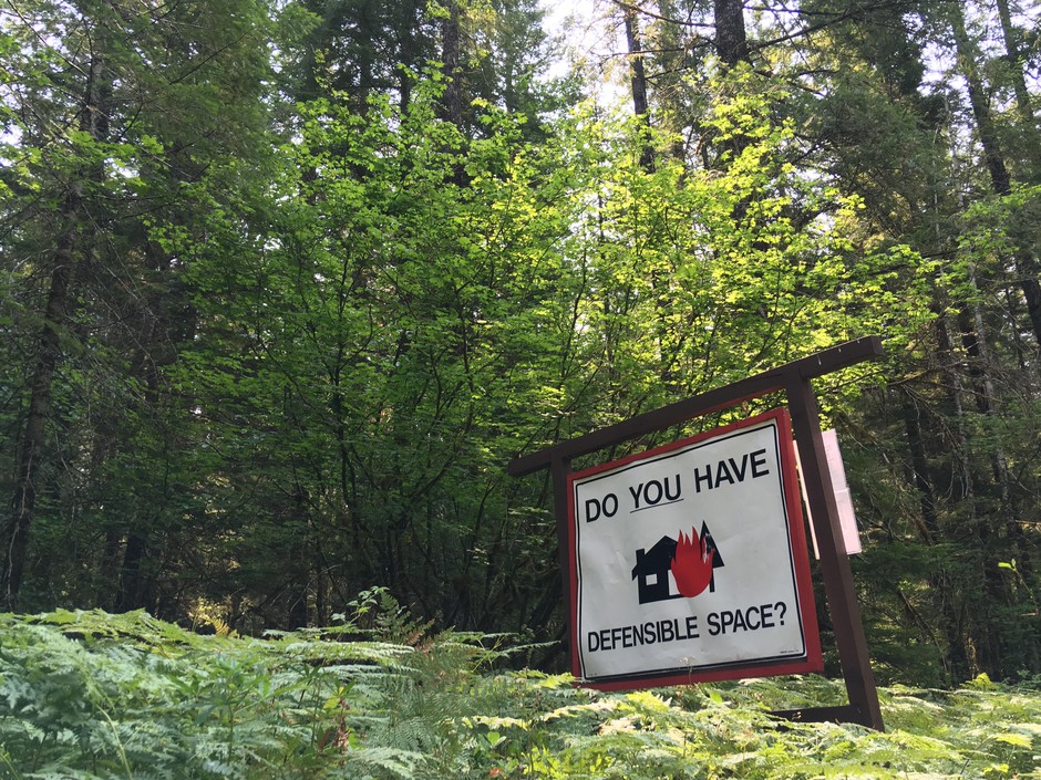 A roadside reminder reminder in a forest reads “Do you have defensible space?” with an illustration of a house engulfed in flames.