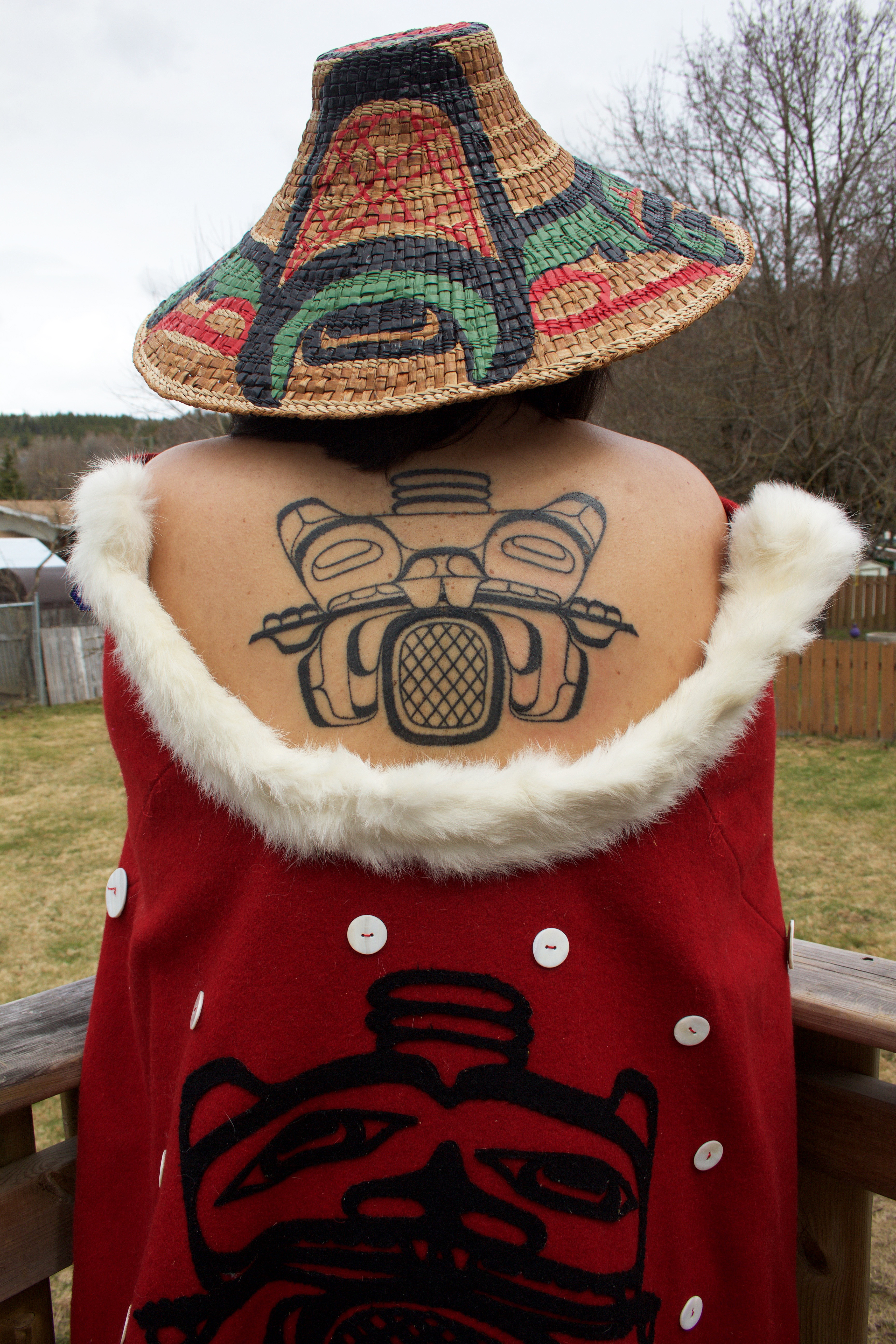 Person stands with back to camera, a tattoo depicted on her back. The same drawing is seen on something she is wearing around her as well. She is wearing a woven hat.