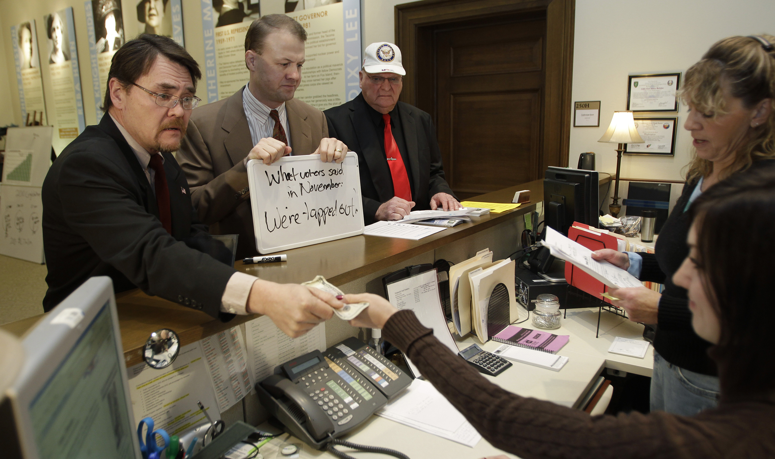 Tim Eyman stands at a reception desk next to a man paying a $5 filing fee