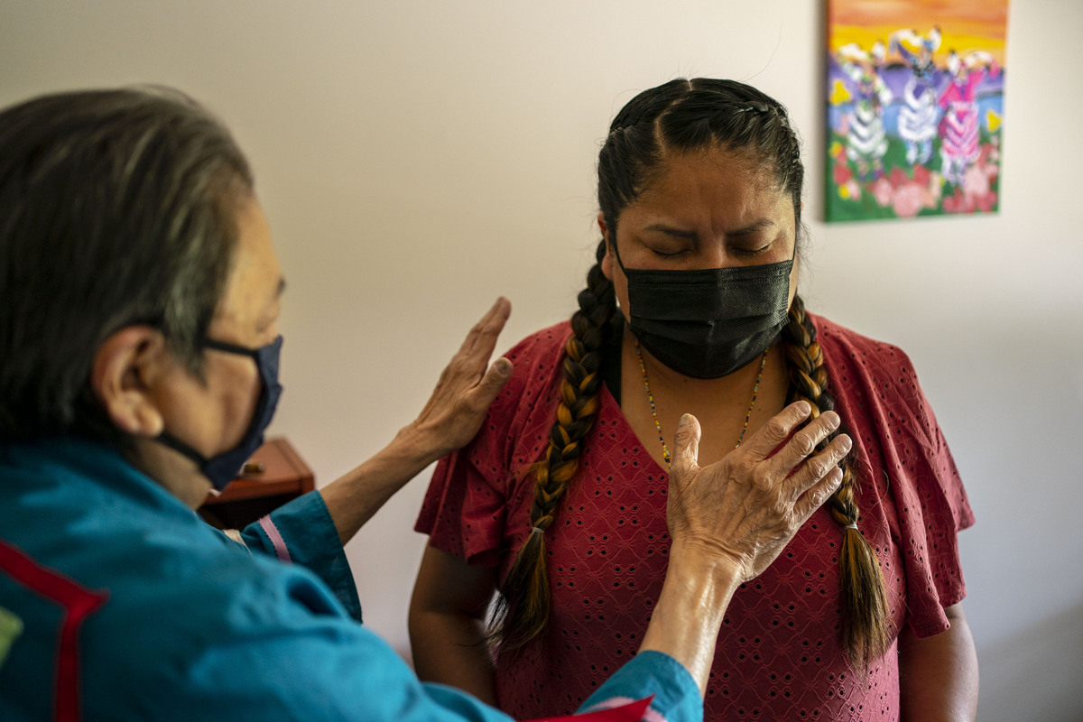 A woman in a face mask stands with her eyes closed as an elder stands over her