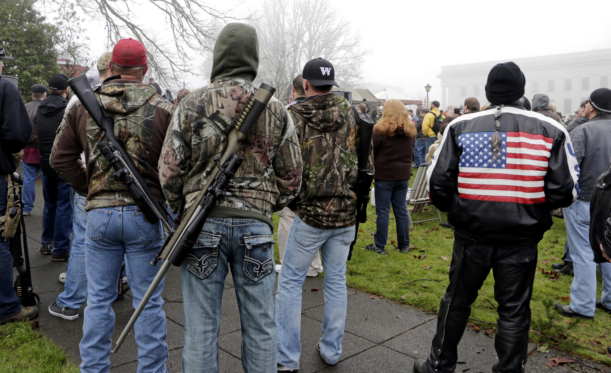 On a foggy day, a crowd of people faces away from the camera. Two men standing in the left side of the frame have large rifles slung diagonally across their backs. 