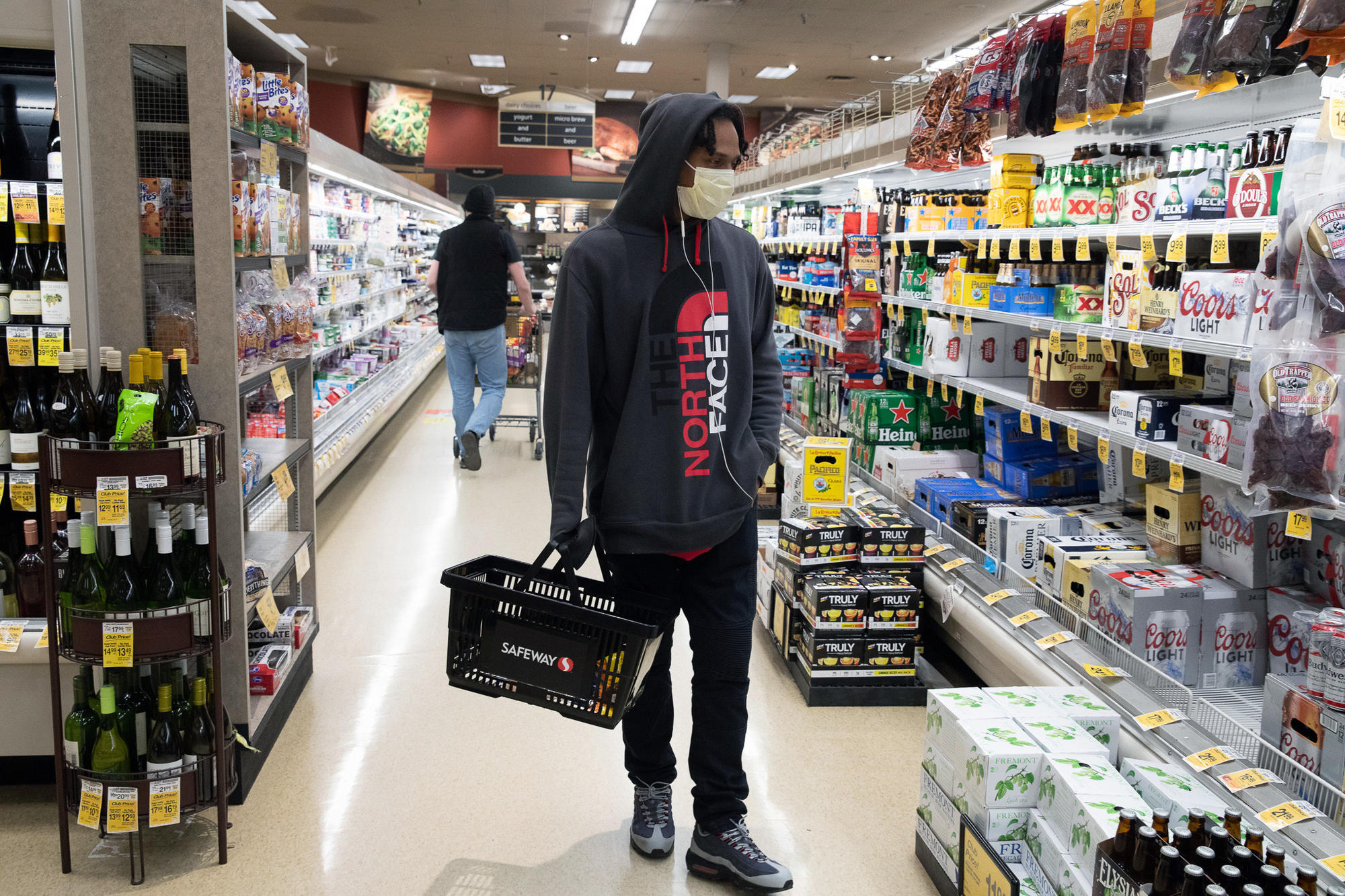 A man wearing a face mask walks down a grocery store aisle with a shopping basket in his hand