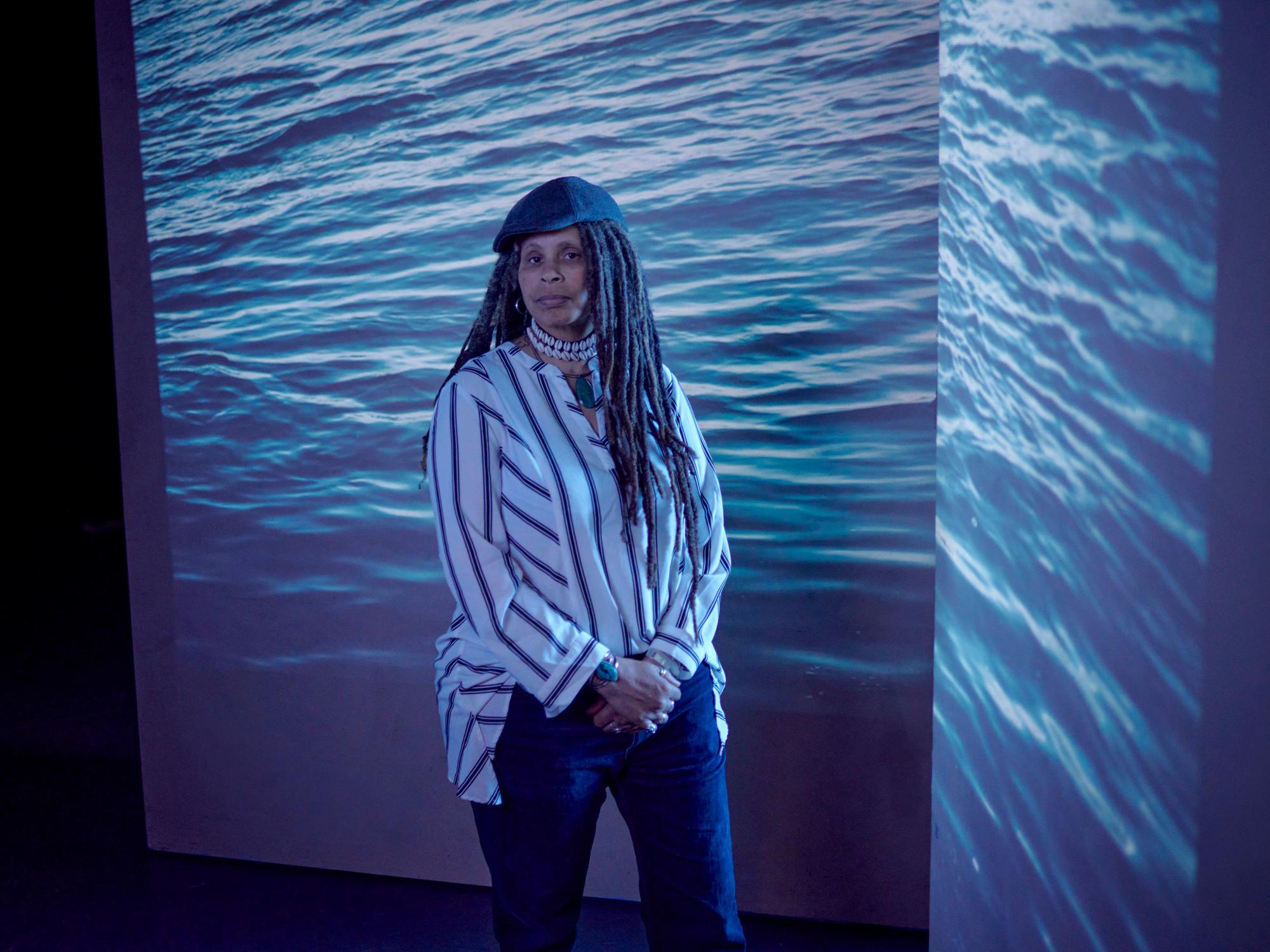 Portrait of Keith in a striped shirt, hat and dreadlocks with a photo of the ocean projected in the background