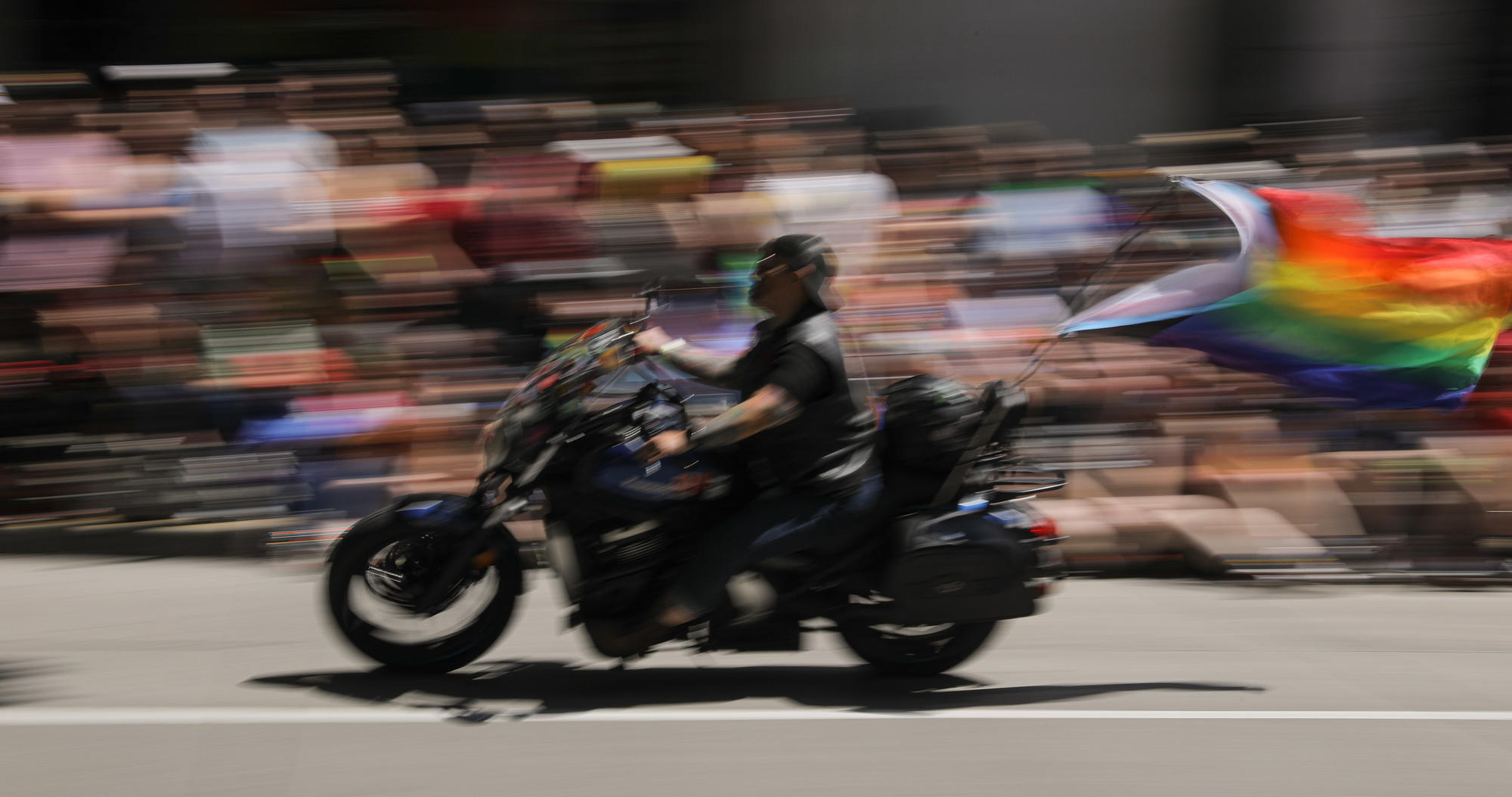 A slow shutter panning shot of a rider on a motorcycle with a rainbow Pride flag blowing behind her